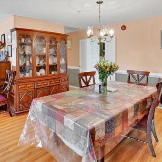 A cozy dining room with a wooden table covered with a colorful tablecloth, surrounded by chairs and illuminated by a chandelier. A china cabinet is seen in the background on a hardwood floor.