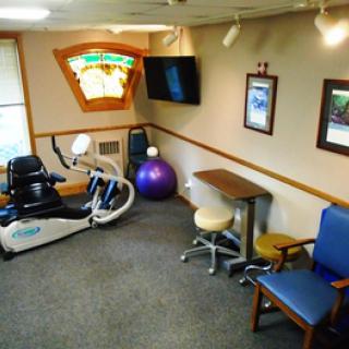 A small exercise room featuring a stationary bike and an exercise ball, with a television mounted on the wall and seating available.