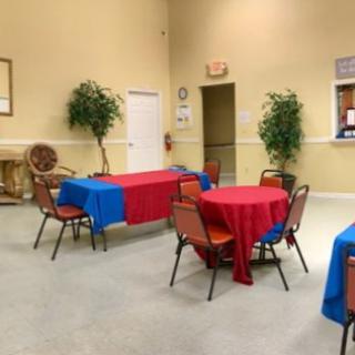 A decorated event hall with tables and chairs covered in blue and red tablecloths, surrounded by potted plants.