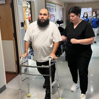 A patient uses a walker while being assisted by a caregiver in a hospital setting.