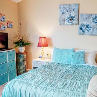 A cozy bedroom featuring a blue color scheme with decorative pillows, artwork, and a bedside lamp.
