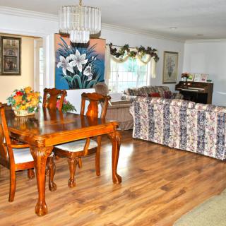 A cozy dining and living area featuring a polished wooden dining table, floral artwork, and comfortable seating with bright lighting.