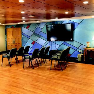 A modern meeting room with rows of chairs facing a television mounted on an artistic wall, featuring a wooden floor and ceiling lights.