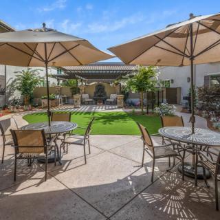 A sunny outdoor patio area with tables, chairs, and large umbrellas providing shade, surrounded by greenery.