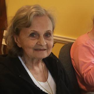 An elderly woman smiling while sitting indoors in casual clothing.