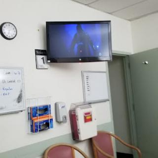 A view of a hospital waiting room featuring a wall-mounted TV, chairs, and a clock.