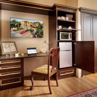 A cozy home office setup with wooden furniture, featuring a desk, chair, laptop, shelving with a microwave and small refrigerator.