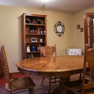 A cozy dining room with a wooden table and chairs, a bookshelf in the corner, and a wall clock.
