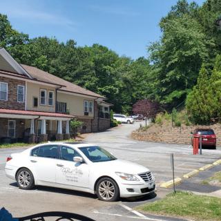 A white car parked in a lot near residential buildings surrounded by trees on a sunny day.