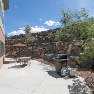 A sunny backyard patio with a table, chairs, and a grill set against a rocky landscaped backdrop.