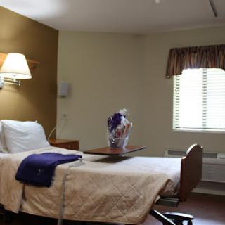 A serene hospital room featuring a bed, bedside lamp, window with curtains, and a small table with a decorative vase.