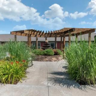 A scenic pathway leads to a wooden pergola surrounded by lush greenery under a bright blue sky.