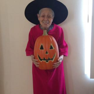 A person dressed in a witch costume holding a carved pumpkin indoors, smiling at the camera.