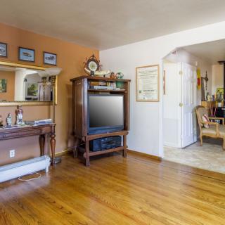 A cozy living room with wooden flooring, featuring a television and elegant furniture, leading to an open archway into another area.