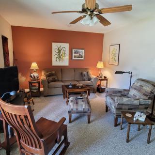 A cozy living room with a sofa, armchair, ceiling fan, and artwork on the walls.