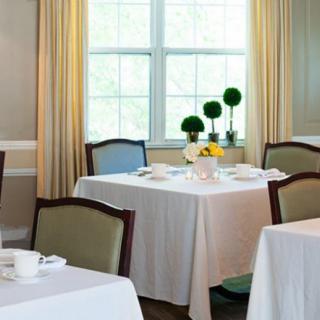 A neatly arranged dining area with tables covered in white tablecloths, decorated with plants and set with tableware near large windows.