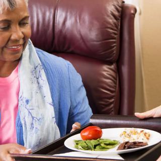 An elderly woman receives a meal on a tray, indicating assistance and care.