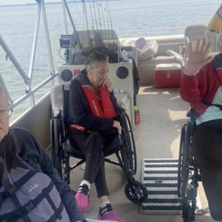 A group of elderly individuals in wheelchairs enjoying a boat ride on a lake, surrounded by fishing rods and wearing life jackets.