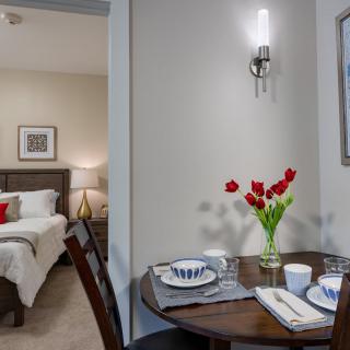 A cozy home interior featuring a dining table set with plates and a vase of red flowers, adjacent to a warmly lit bedroom.