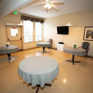 A well-lit room with round tables covered with cloth, chairs, a television, and a ceiling fan.