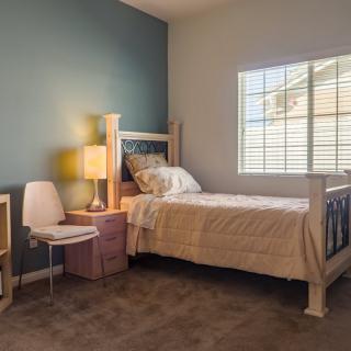 A cozy bedroom featuring a single bed, a bedside lamp on a small table, and a chair near the window.