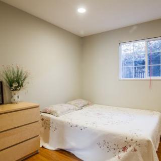 A minimalist bedroom with a simple bed, wooden dresser, and a plant by the window.