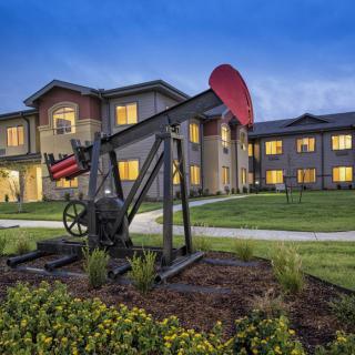 An oil pumpjack in front of a lit residential building during the evening.