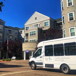 A white shuttle bus parked in front of a multi-story residential building on a clear day.