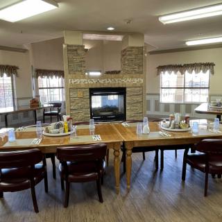 A cozy dining room with tables set for a meal, featuring a central fireplace and large windows.