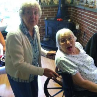 Two elderly women sharing a moment indoors, one seated in a wheelchair.