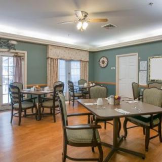 A dining room with multiple tables and chairs arranged on a hardwood floor, featuring a ceiling fan and large windows with curtains.