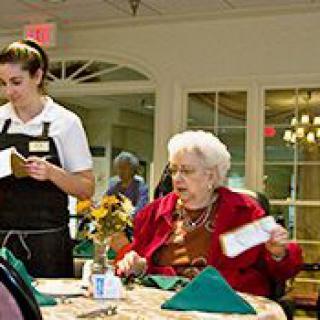 An elderly woman seated at a dining table interacting with a waitress in a restaurant setting.