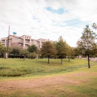 A scenic view of a park with buildings in the background, surrounded by trees and green grass under a partly cloudy sky.