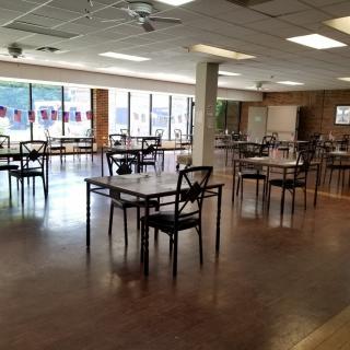 An empty dining area with tables and chairs, featuring large windows and a hardwood floor.