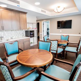 A modern dining area with wooden tables and blue upholstered chairs, featuring a kitchenette and a wall-mounted television.