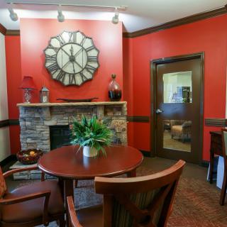 A cozy office meeting room with red walls, featuring a round table, chairs, a large decorative clock above a stone fireplace, and a potted plant.