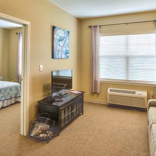 A cozy living room with a TV stand, sofa, and a view into the adjacent bedroom. The space is softly lit through a large window with curtains.