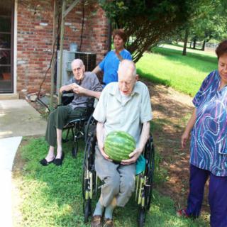 A group of elderly people, one holding a watermelon, enjoys time outdoors with caregivers.