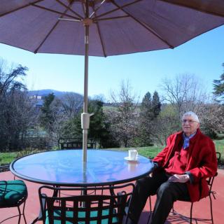 An elderly person sitting at a patio table under an umbrella, enjoying a sunny day with a scenic view in the background.
