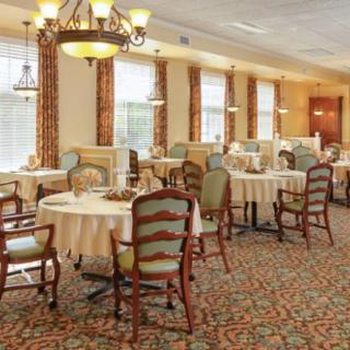 A spacious and elegantly decorated dining room with round tables and comfortable chairs, featuring chandeliers and a carpeted floor.