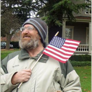A bearded man wearing a beanie and jacket is holding an American flag while standing outdoors in front of a house.