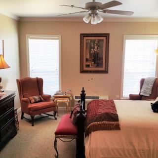 A cozy and well-furnished bedroom featuring a bed with pillows, two armchairs, a dresser, and a ceiling fan, all complemented by soft lighting and artwork.