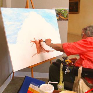 An artist in a wheelchair working on a painting with vibrant colors on a canvas inside a studio.