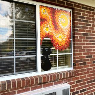 A bright artwork featuring a sun pattern is displayed in a window, while a cat silhouette peeks through the blinds.