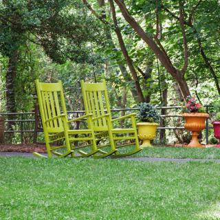 A serene garden setting with two bright yellow rocking chairs on a lush green lawn surrounded by trees and colorful potted plants.