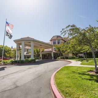 A well-maintained building entrance with a circular driveway and flags, surrounded by lush trees and landscaping on a sunny day.