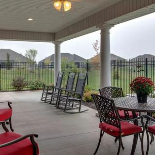A cozy covered patio with rocking chairs and a table with red-cushioned chairs, overlooking a fenced lawn.