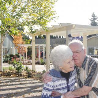 An elderly couple embraces warmly outdoors on a sunny day in a garden setting.