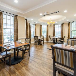 A cozy dining area with neatly arranged tables and chairs, featuring large windows and a central chandelier.