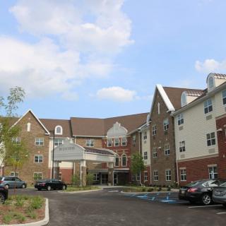 A multi-story residential building with a parking lot and cars in front, surrounded by trees under a partly cloudy sky.